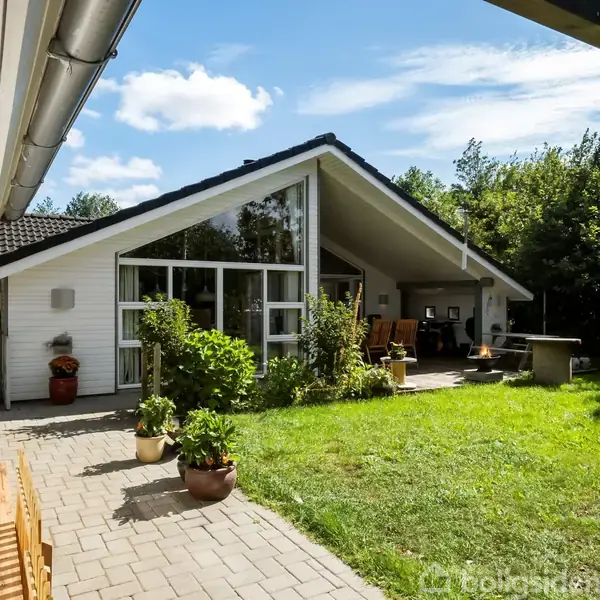 Hus med store glasvinduer står fredfyldt i solskinsvejr, omkranset af grønne planter og en velholdt græsplæne. Nogle havemøbler er placeret på en terrasse under husets udhæng.