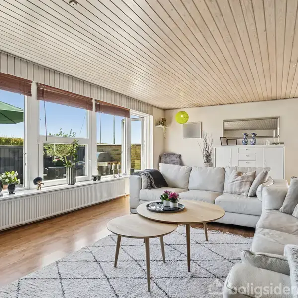 En stue med en hvid sofa og to runde sofaborde står i et lyst rum med store vinduer. Udenfor ses en grøn have med parasol.