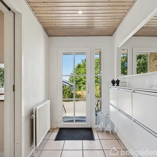 En lys entré med klinkegulv og træloft, hvor en hvid skoskab står ved væggen. Glasdøre fører ud til en terrasse med udsigt til grønne omgivelser.