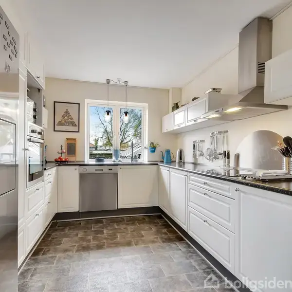 Køkken med hvide skabe og sort bordplade, fyldt med køkkenredskaber. Stor stål-køleskab ved siden af indbyggede ovne. Vindue mod udendørs, der slipper lys ind. Vægge dekoreret med billeder.