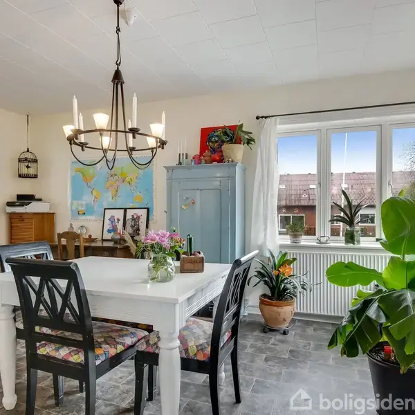 Et spisebord med blomster og stole står under en lysekrone. Ved siden af er et blåt skab. Vægkort og planter pryder rummet, som har store vinduer med udsigt.
