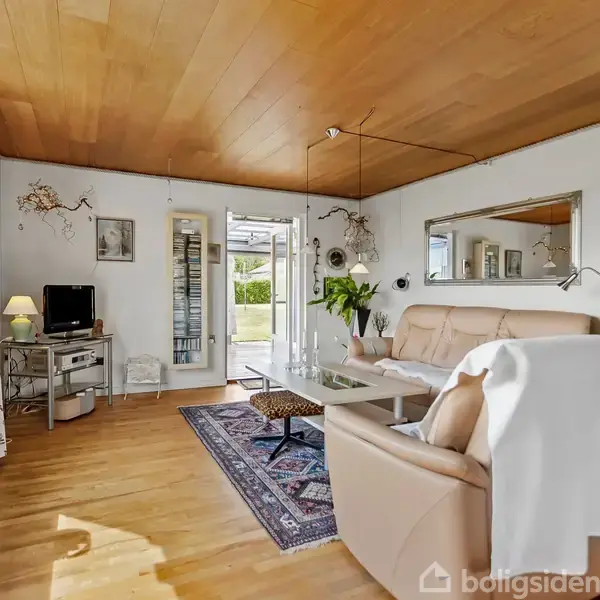 Sofagruppe med beige farver står på et trægulv, flankeret af et sofabord. Rummet har store vinduer med lysindfald, indrettet med planter og en lille fjernsynsbord ved siden af.