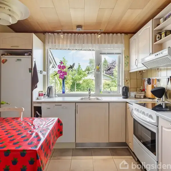 Et køkkenbord dækket med en rød dug står ved siden af et vindue. Bordet har en blomstervase. Baggrunden viser køkkenskabe, en ovn, og udsigt til en have.