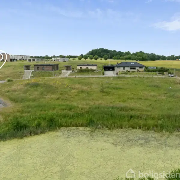 En vej løber gennem et boligområde, hvor et kortikon angiver en specifik placering. Græsbevokset område og huse danner baggrunden med åbne marker og trælunde i det fjerne.