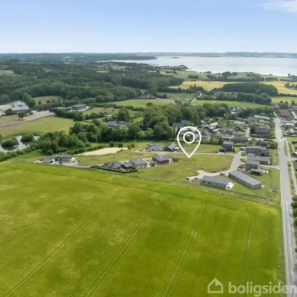 Huse ligger spredt i et grønt landsbynabo, nær en større vej. En pin-ikon angiver en bestemt placering. I baggrunden ses skov og vand.