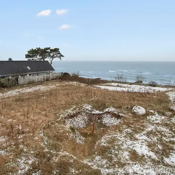 Et snedækket græsareal med en lavning i midten strækker sig ned mod havet. Til venstre står et hus med et skråt tag, omgivet af et hegn.