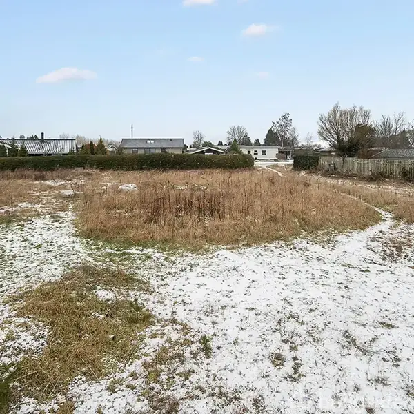En snedækket grund med en cirkulær græsbevokset område i midten. Omgivet af hegn og huse i en rolig beboelsesområde under en klar blå himmel.