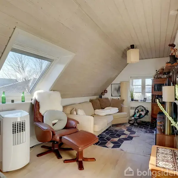 Et hyggeligt stueområde med skrå loft, hvor en lænestol og en sofa er placeret. Rummet indeholder også grønne planter, en luftrenser og hælder store vinduer lys ind i rummet.