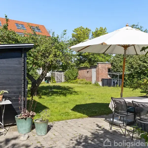 Terrasse med havemøbler og parasol står på flisebelagt område ved et hus. I baggrunden ses en grøn have med træer og en rød murbygning. Solrigt vejr.