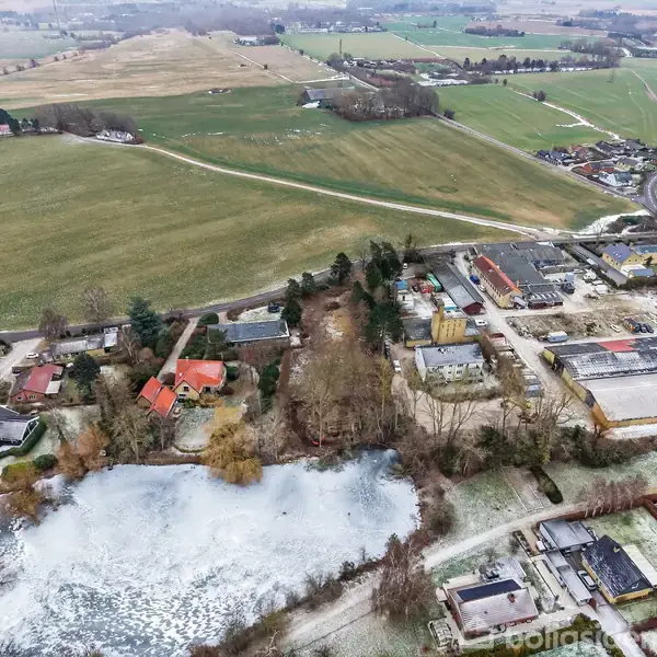 Landsbyområde set fra luften med marker i baggrunden. I forgrunden er der et frossent vandhul omgivet af træer og huse. Veje forbinder forskellige afsnit af området.