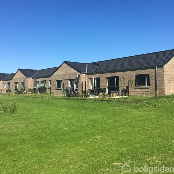 Række af enfamiliehuse med muret facade og sorte tage, stående på en velplejet, grøn græsplæne under en klar blå himmel. Ingen tekst er synlig i billedet.