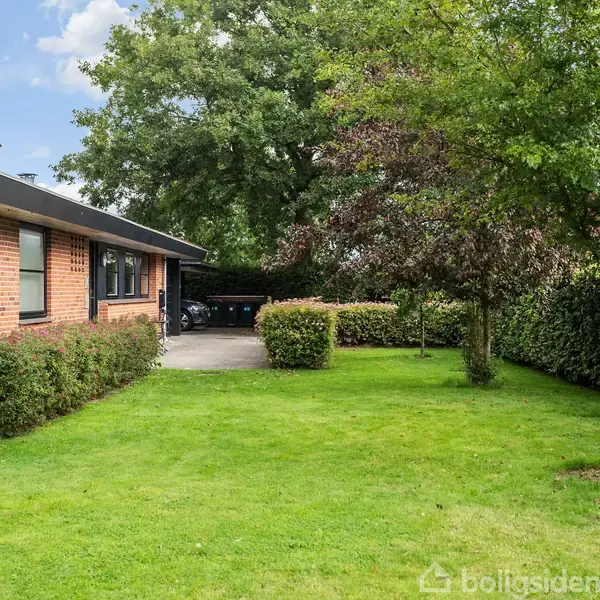 En rød murstensbygning står stille ved siden af en velplejet græsplæne og omgivet af træer og buske; en indkørsel ses i baggrunden under et stort træ.