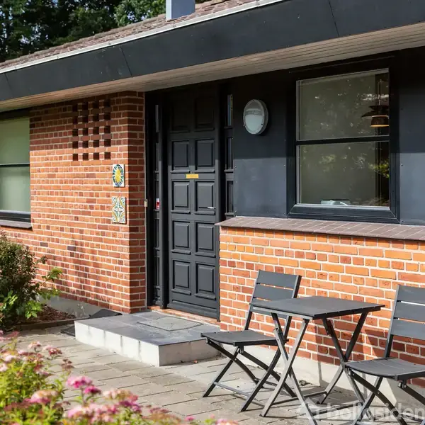 Et rødt murstenshus med en sort dør og vinduer. Foran huset står to sorte stole og et bord på en flisebelagt terrasse. Blomsterbed fyldt med lyserøde blomster langs kanten.