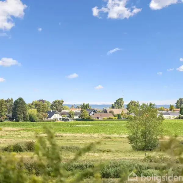 Grønne marker strækker sig foran en række huse, der ses i baggrunden under en klar blå himmel med spredte hvide skyer.
