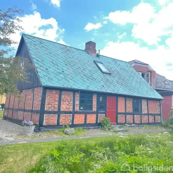 Hus af mursten og bindingsværk med grønt tag og rød dør, står i en have med græs og træer under en blå, skyfuld himmel.