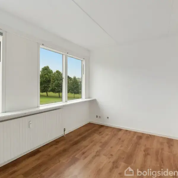 Et lyst rum med trægulve har store vinduer med udsigt til grønne træer og en græsplæne udenfor. Rummet fremstår tomt og nyistandsat.