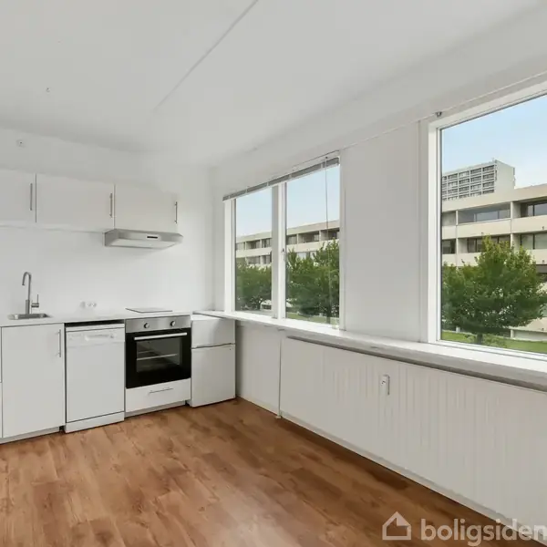 Et køkken med hvide skabe og en ovn står i et rum med trægulv. Store vinduer viser en udsigt til en bygning og træer udenfor.