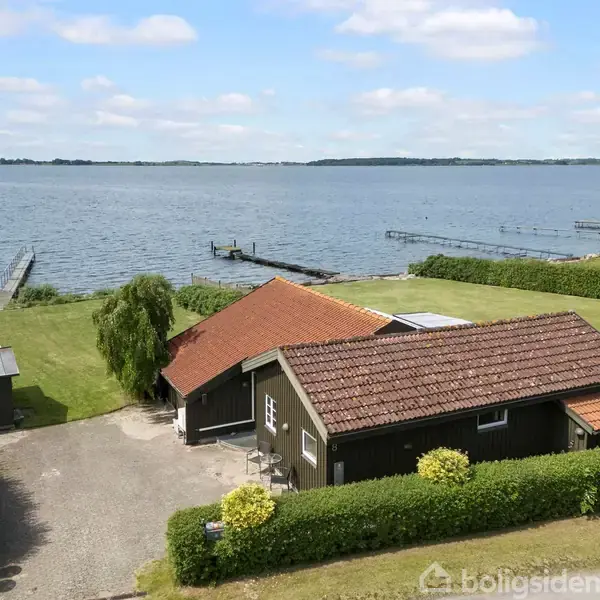 Et træhus med rødt tegltag ligger ved en sø. Haven er omgivet af en hæk, og en bådbro fører ud i vandet. Skyfri himmel.