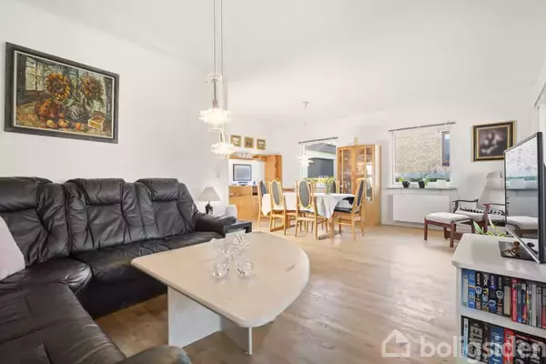 Sort lædersofa og sofabord står i stue, hvor spisebord med stole ses i baggrunden. Væggene har billeder, og der er et skab ved vinduet. Der er lamper, et tv og bogreol.