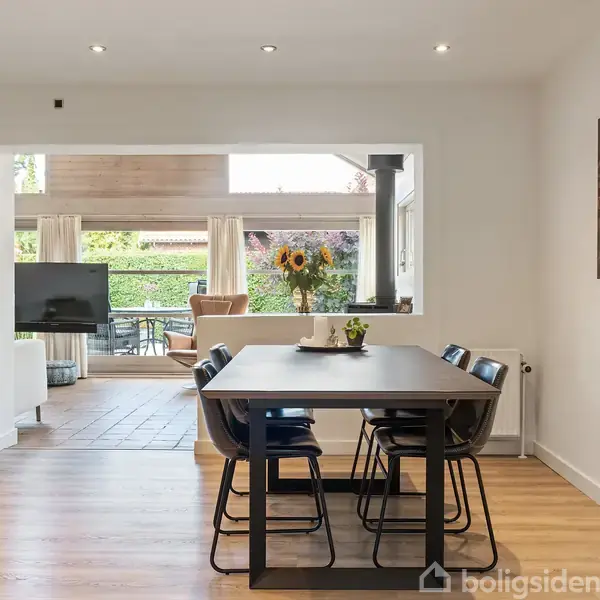 Et moderne spisebord med sort bordplade står i et lyst rum med fire sorte stole omkring det. Til venstre er en grøn plante, og i baggrunden ses en stue med tv.