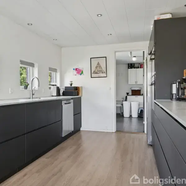 Sort moderne køkken med mørke skabe og lyst trægulv, åbner ud mod stueområde gennem en hvid dør. Tre små vinduer bringer lys ind i rummet.