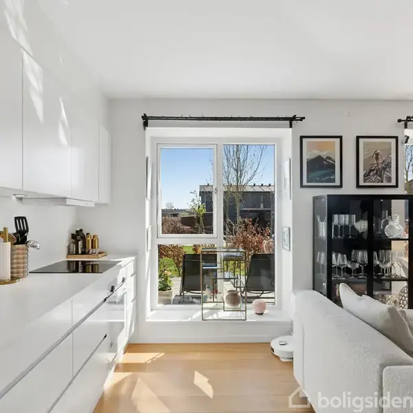 Udendørs flisebelagt terrasse med to liggestole foran åbne glasdøre, der fører ind til en stue. Omgivelserne omfatter en murstensbygning og moderne lejlighedskomplekser i baggrunden.