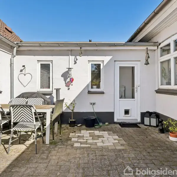 Terrassebord med stole står på en flisebelagt gårdsplads. Hvide mure omfavner området, dekoreret med lamper og hjertepynt. En dør og vinduer viser indgangen til huset.