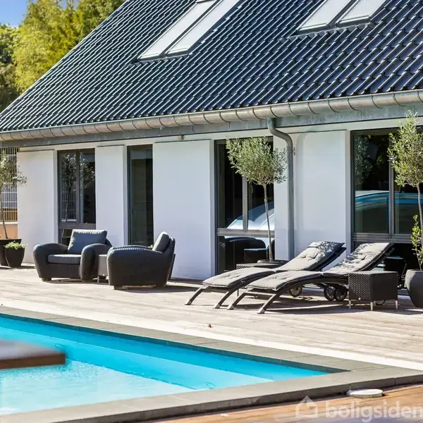 En moderne terrasse med solsenge og stole står ved siden af en swimmingpool. I baggrunden ses et hvidt hus med store vinduer og et mørkt tag.