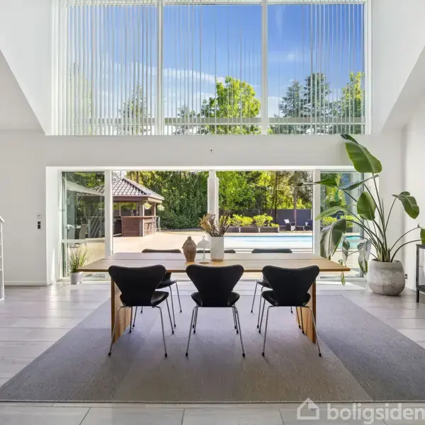 Et moderne spisebord med tre stole står foran store vinduer, der åbner ud til en have med pool. Et stort kunstværk hænger på væggen, og der er planter omkring.