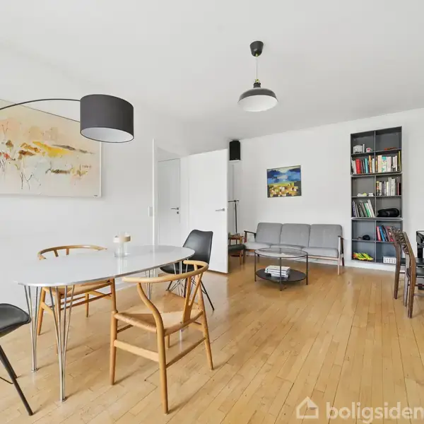 Et moderne stueinteriør med lyse træplankegulve og hvide vægge. Der er et bord med stole, en sofa ved en væg med hylder og et klaver med lampe.