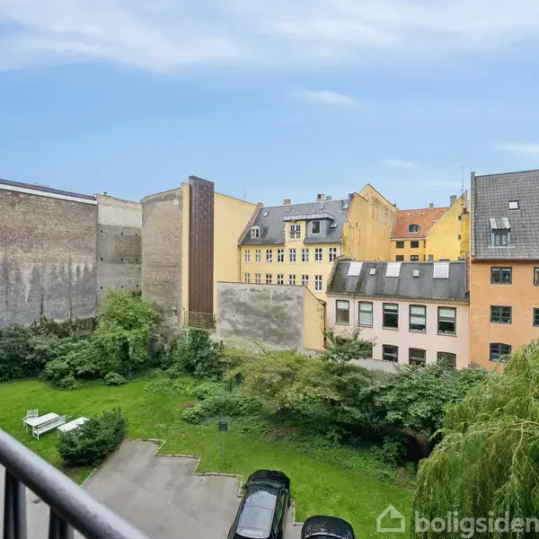 Udsigt fra en altan over en baggård med parkeringsplads og grønne områder; omgivet af ældre bygninger med gule og grå facader, og blå himmel i baggrunden.