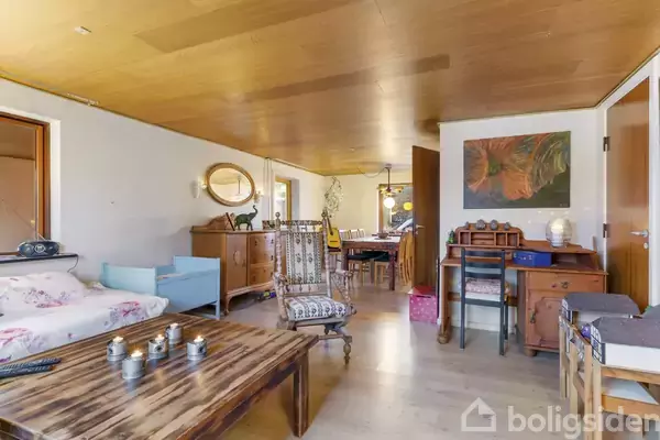 En stue med en træbord i centrum; omkring det er en sofa, stole og skabe. Væggene har billeder og spejl. I baggrunden ses en spisestue med lamper.