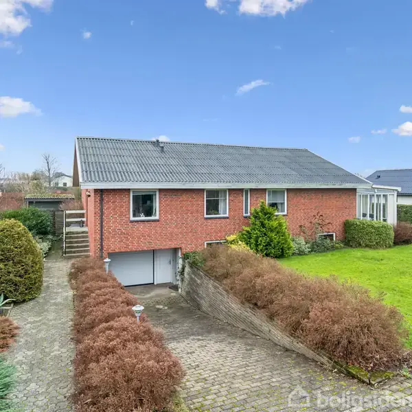 Rødt murstenshus med mørkt tag, beliggende ved en velplejet have og flisebelagt indkørsel. Flere buske og grønne områder omkranser huset under en klar blå himmel med spredte skyer.