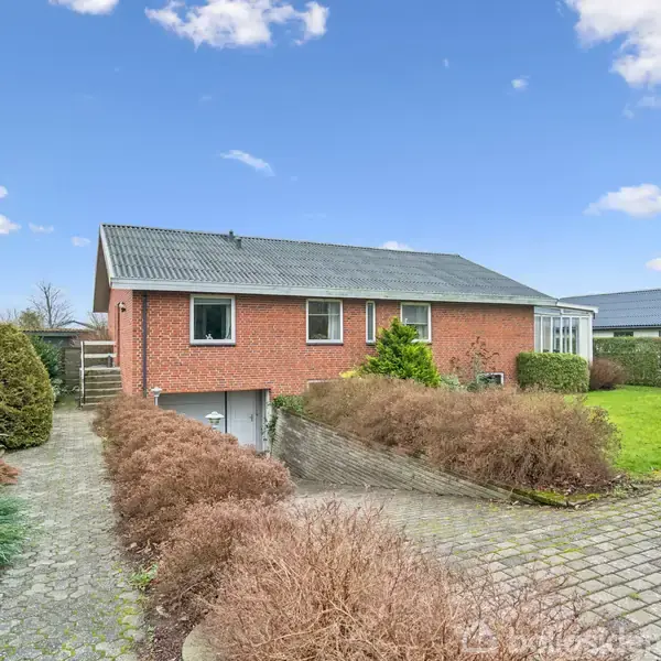 Rødt murstenshus står stille med garageadgang nedenfor, omgivet af grønne buske og græsplæne, under en blå himmel med spredte skyer.