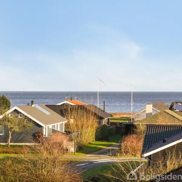 Huse står stille i en kystby med havudsigt i baggrunden. Omkringliggende træer og buske rammer scenen i sollys.