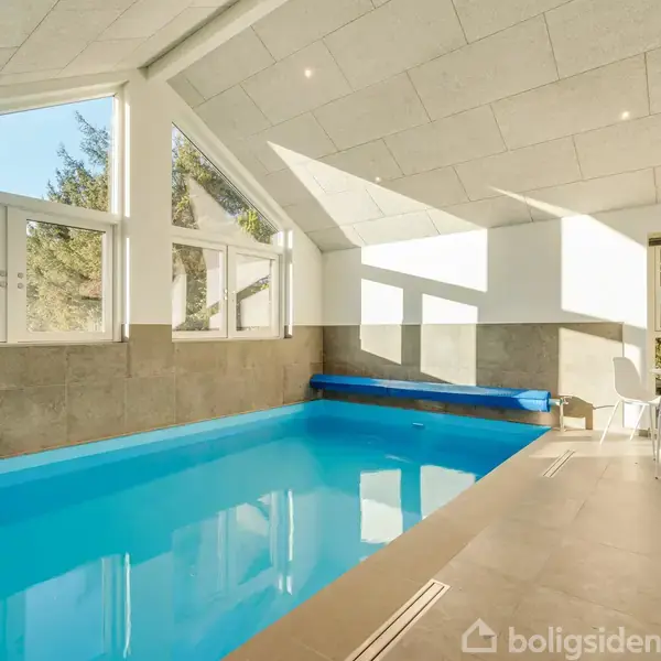 Indendørs swimmingpool med blåt vand står stille i et lyst rum med store vinduer. Ved siden af er et lille bord med fire hvide stole. Udenfor ses træer og grønt.