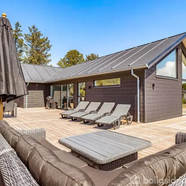 Et moderne sommerhus med sort træfacade har en stor terrasse, hvor solsenge og møbler er stillet op under en parasol. Omgivet af træer under en klar blå himmel.