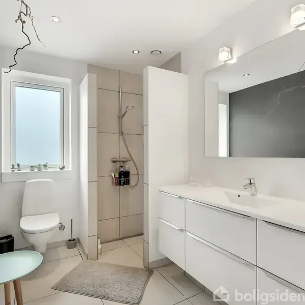Et funktionelt badeværelse indeholder et toilet, dobbelthåndvask med spejl og bruser i et flisebeklædt rum. Et lille, rundt bord står ved siden af toilettet.