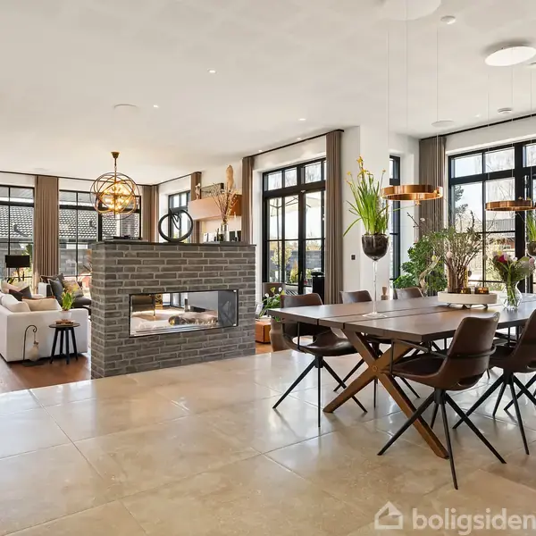 Et stort, moderne rum indeholder en elegant spisebordsgruppe med otte stole. Ved siden af, en åben stue med sofaer og en indbygget pejs i midten af rummet. Store vinduer giver naturligt lys.