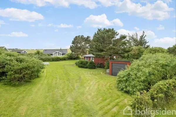 Grøn græsplæne strækker sig foran en rødmalet træbygning omgivet af buske og træer. I baggrunden ses flere huse under en blå himmel med spredte skyer.