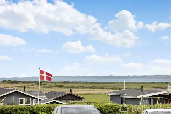 Dansk flag vajer foran kystnære sommerhuse. Foran parkeres en bil, mens baggrunden viser en bred udsigt over vand og himmel med skyer.