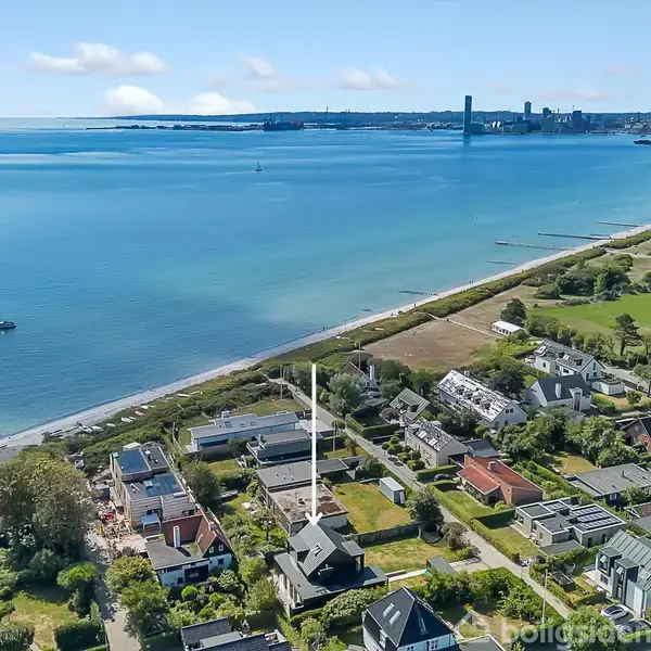 Kystnært boligområde med moderne huse ligger langs en strand. Havet strækker sig til horisonten. I baggrunden ses en by med industri og grønne områder.