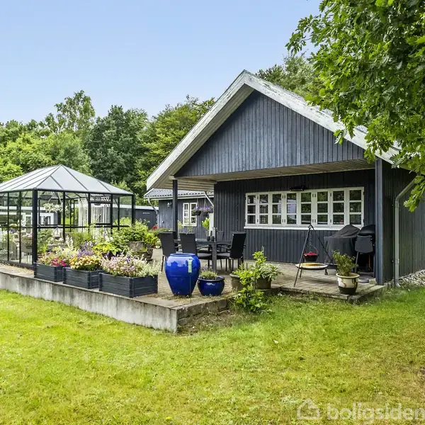 Et sort træhus med hvide vinduer står ved siden af et drivhus fyldt med blomsterplanter. Omkring bygningerne er der en grøn have med frodige træer.
