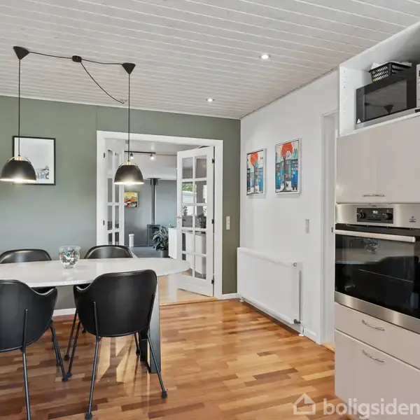 Et spisebord med sort stole i et køkken-alrum, omgivet af hvide vægge og en grøn bagvæg. Der er moderne belysning over bordet og trælaminat på gulvet.