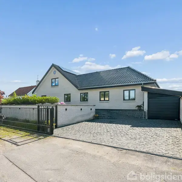 En moderne villa med gråt tag og lys facade står roligt i en forstad. Foran huset er der en stenbelagt indkørsel og en lukket garage. Solen skinner klart.