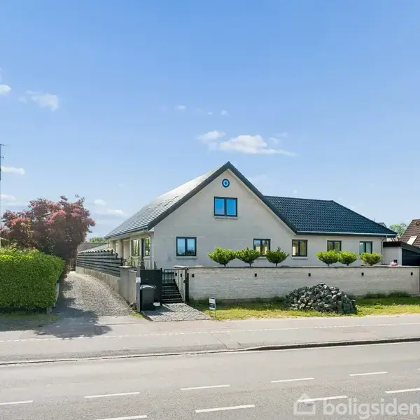 Et stort, moderne hus med sort tag står ved en vej. Huset har flere vinduer og en velplejet have indenfor en lav hvid mur. Solrig dag med få skyer på himlen.