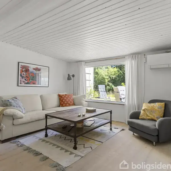 En stue med en off-white sofa og en grå lænestol står omkring et træsofabord. Store vinduer viser en grøn udendørs have. Mønstret tæppe og farverige puder tilføjer hygge.