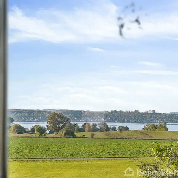 Et vindue tilbyder udsigt over en grøn mark og en fjord i baggrunden under en blå himmel med spredte skyer.