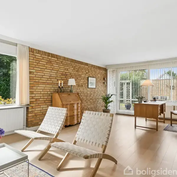 En moderne bungalow med fladt tag står stille omgivet af buske og træer i et roligt nabolag. En gadelampe står til venstre, og der ses en lys himmel.