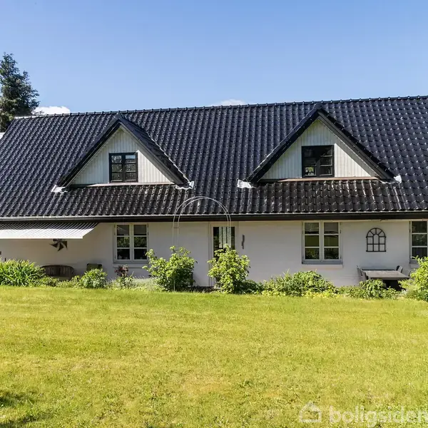 En hvid, to-etagers villa med et sort sadeltag står i en velholdt have. To kviste pryder taget, mens grønne buske omgiver huset. Blå himmel i baggrunden.